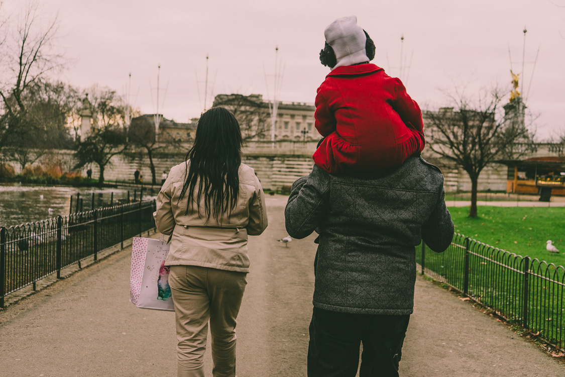 Candid photograph of a family walking through St James’s Park in London, seen from behind. A man carries a child wearing a bright red coat on his shoulders as they stroll together.