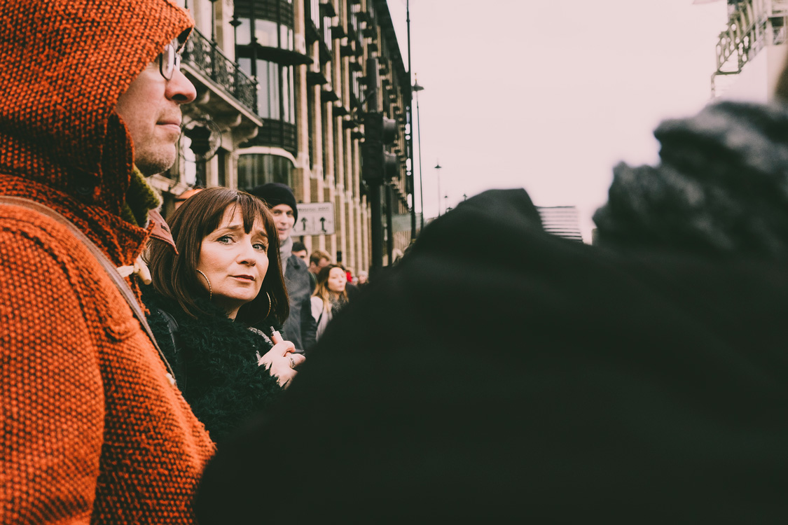 Candid photograph of people waiting to cross a street in London, viewed over the shoulder of one person. A man in an orange coat stands nearby, while a woman looks directly at the camera with a soft, inquisitive smile.