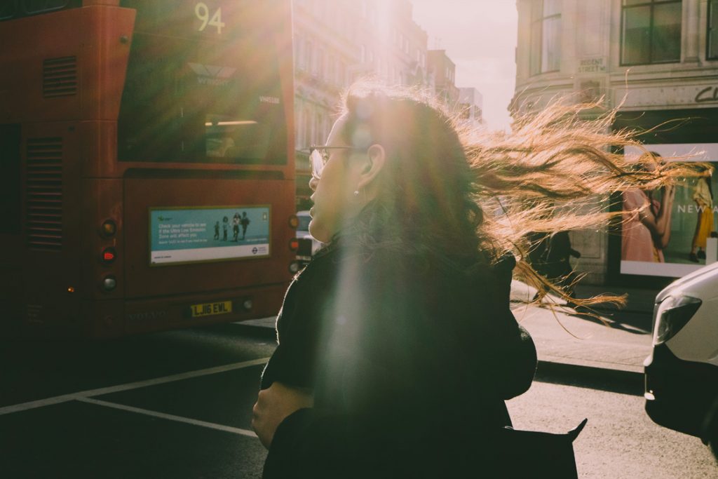 A candid photo of a woman walking on a busy street, backlit by sun rays that illuminate her blowing hair in dramatic sunlight.