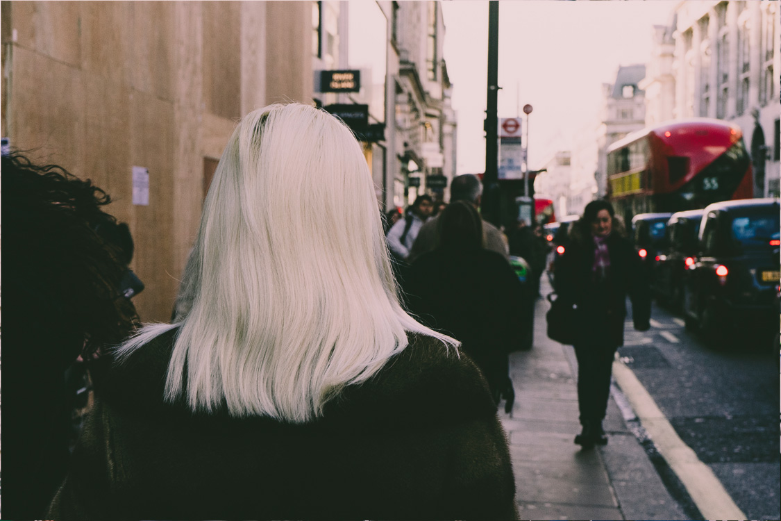 A woman with striking white-blonde hair walking ahead, photographed from behind; busy street with cabs, red buses, and people to the right.