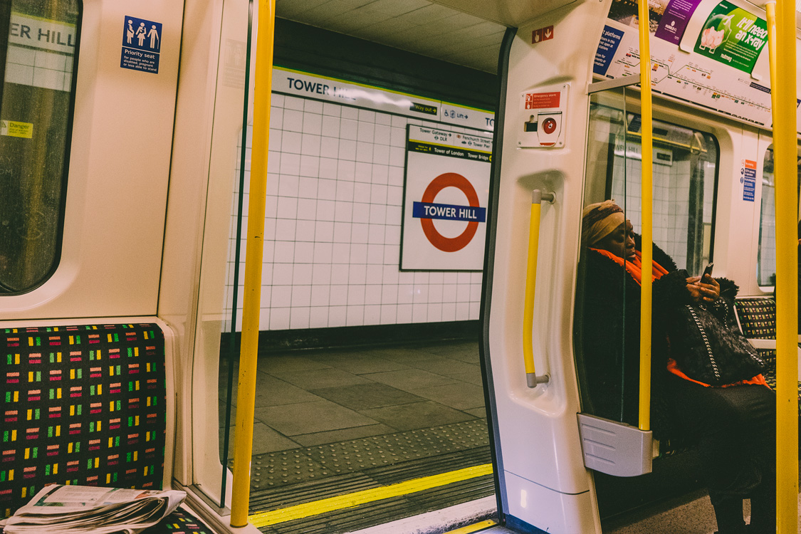 Candid photo from inside a Tube train at Tower Hill station with open door, showing the Underground logo, a seated woman, and an abandoned newspaper.