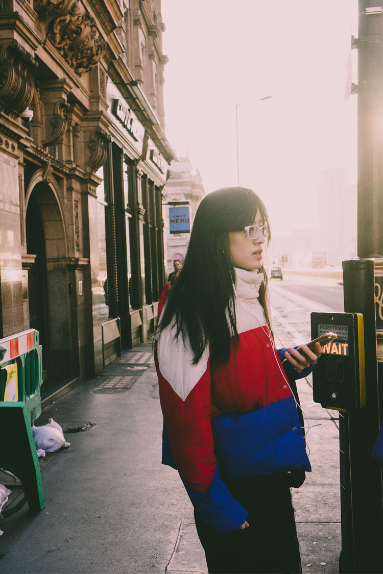 Candid street portrait of a woman in red, white, and blue jacket waiting to cross foggy London street, city elements in background.
